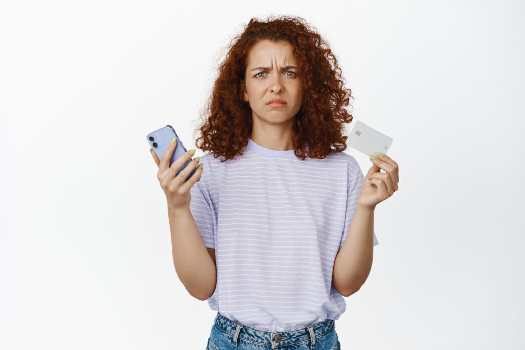 Disappoined, confused redhead girl holding credit card and mobile phone, frowning puzzled, dont understand, standing over white background